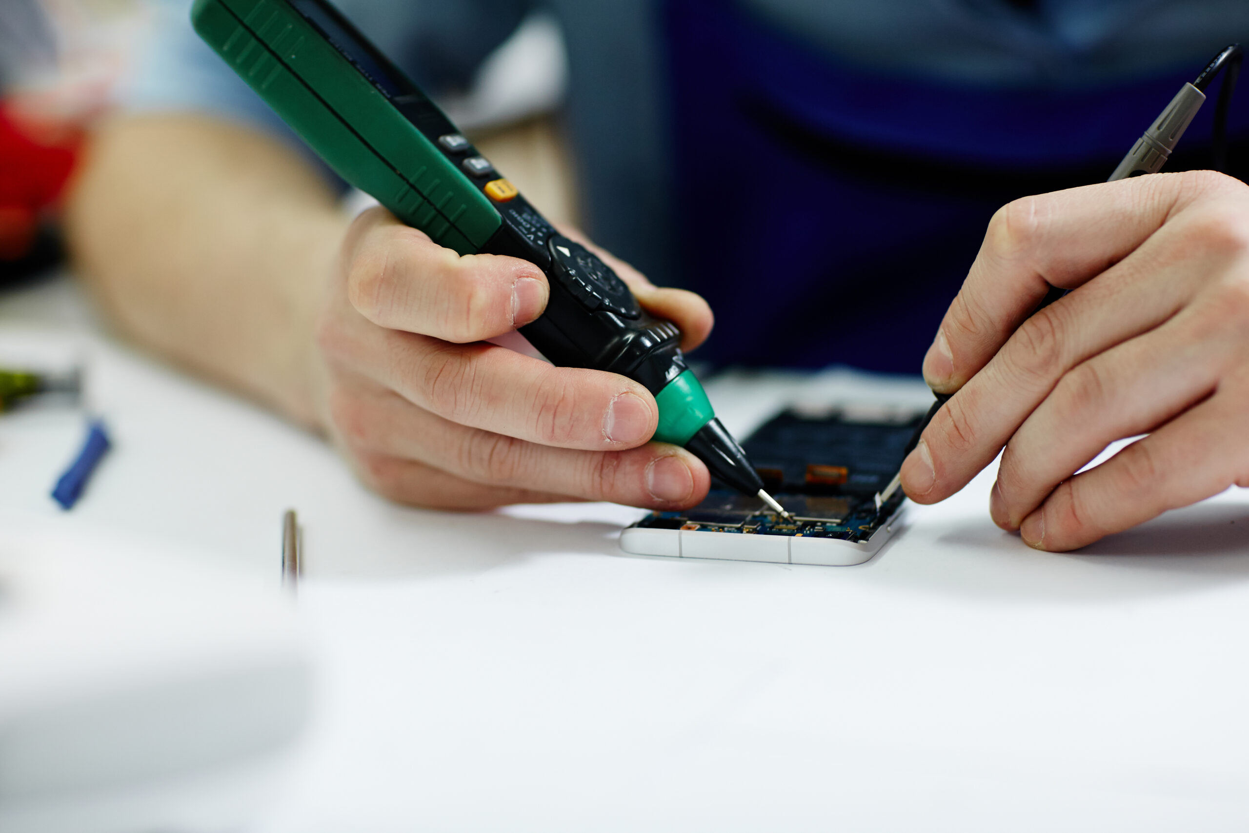 Close-up van handen die bezig zijn met het demonteren van een printplaat in een mobiele telefoon en het controleren van de elektrische stroom met een multimeter.